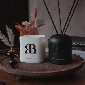 May include: Two black and white glass containers with the letters "RB" and the text "bloom boutique" on them. The container on the left is a scented candle and the container on the right is an aromatherapy diffuser. The diffuser has black sticks in it. The containers are on a wooden tray.