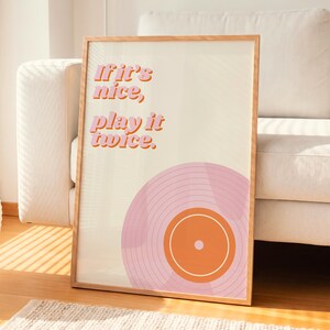 May include: A framed wall art print featuring a light cream background with the text If it's nice, play it twice. in pink and orange letters. Below the text is a graphic illustration of a vinyl record in shades of pink and orange.