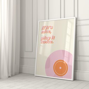 May include: A framed rectangular wall art print featuring a cream background with the pink and orange text "If it's nice, play it twice." Below the text is a graphic illustration of a pink vinyl record with an orange center label and white rings.