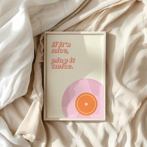 May include: A rectangular framed wall art print featuring a cream background with the orange text "If it's nice, play it twice." and a graphic illustration of a pink vinyl record with an orange center label positioned at the bottom right corner.