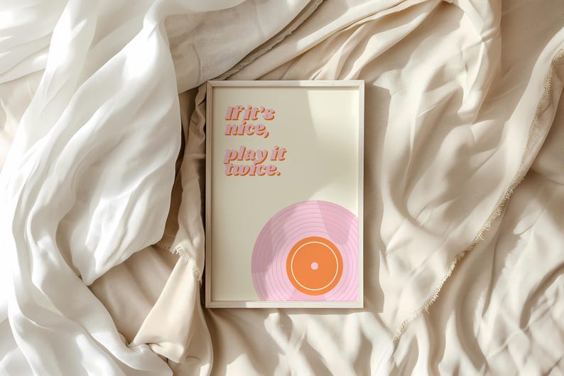 May include: A rectangular framed wall art print featuring a cream background with the orange text "If it's nice, play it twice." and a graphic illustration of a pink vinyl record with an orange center label positioned at the bottom right corner.