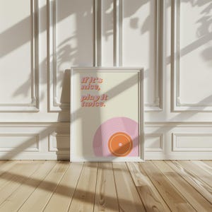 May include: A framed wall art print featuring a cream background with the text 'If it's nice, play it twice.' in a pink and orange font. The bottom right corner displays a stylized graphic of a pink vinyl record with an orange center label.