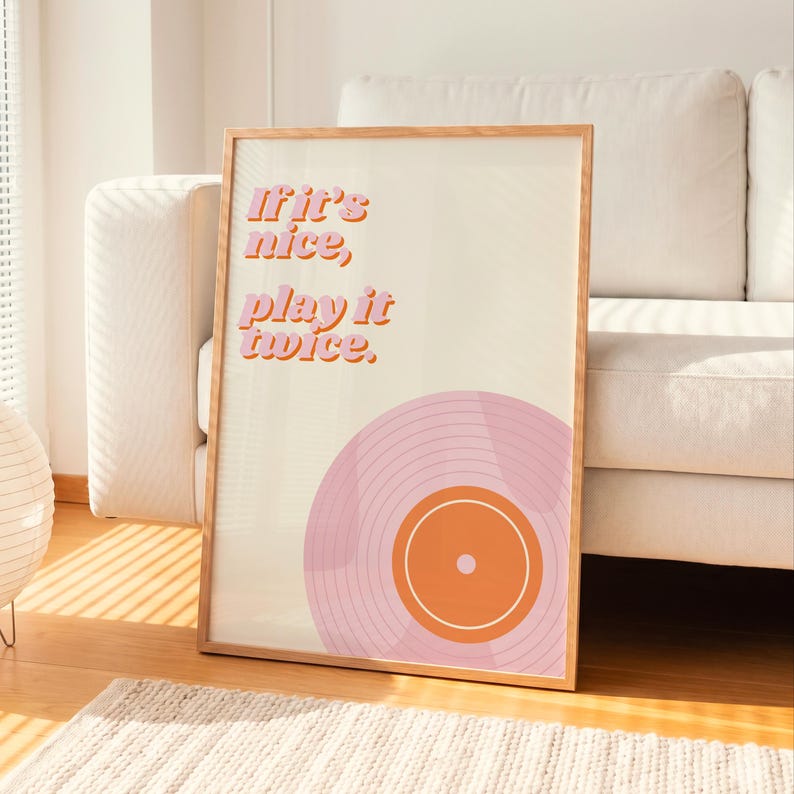 May include: A framed wall art print featuring a light cream background with the text If it's nice, play it twice. in pink and orange letters. Below the text is a graphic illustration of a vinyl record in shades of pink and orange.