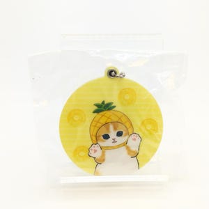 以下が含まれることがあります： パイナップルの帽子をかぶった白い猫が描かれた、丸い黄色のキーホルダー。猫は黄色のパイナップルの輪切りに囲まれています。