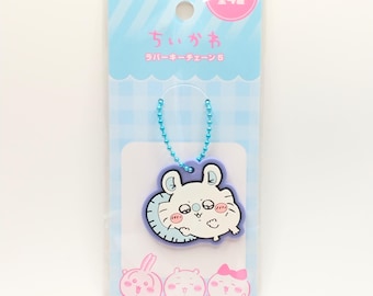 May include: A kawaii-style keychain featuring a white cartoon character with pink cheeks and a blue border. The keychain is attached to a blue ball chain and is packaged on a blue and white striped background. The packaging has Japanese text.