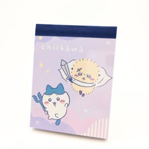 以下が含まれることがあります： 紫色の背景に漫画のキャラクターと「chiikawa」の文字が入ったメモ帳。 一方のキャラクターは青いリボンと三叉槍、もう一方は剣とマントを持っています。 上部のバインディングは濃い青色です。