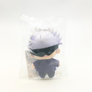 May include: A plush toy of a character with purple hair, wearing a black mask and a purple outfit. The toy is packaged in a clear plastic bag.