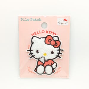 May include: A Hello Kitty iron-on patch on a pink backing. The patch features a white Hello Kitty character with a red bow, wearing a red dress. The backing has the words "Pile Patch" and "Hello Kitty" printed on it.