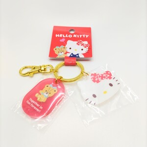 May include: A Hello Kitty keyring with a red oval charm featuring a bear and the text "Together in fun forever!" and a white charm featuring Hello Kitty with a red bow.