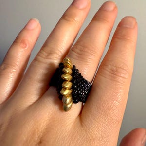 Op de afbeelding: Een zwarte en gouden kralenring met een spiraalvormig ontwerp. De ring wordt gedragen aan een vinger.