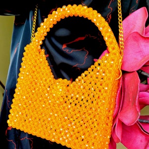 May include: A bright orange beaded handbag with a gold chain strap. The bag is made of small, round beads that are woven together to create a mesh-like design.