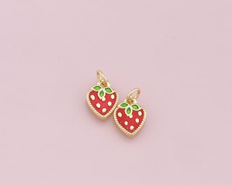 May include: Two gold-colored strawberry charms with red enamel, white dots, and green leaves. The charms are heart-shaped and have a small loop at the top for hanging. The background is a solid light pink.