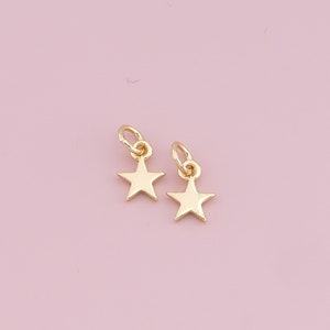 May include: Two small, gold-colored star charms with attached loops, set against a soft pink background. The charms are simple, five-pointed stars, ideal for jewelry making or adding a touch of celestial charm to accessories.