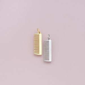 May include: Two gold and silver comb charms. The gold comb charm is on the left and the silver comb charm is on the right. Both charms are on a pink background.