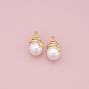 May include: A pair of white pearl earrings with gold snake-like accents. The earrings are on a pink background.