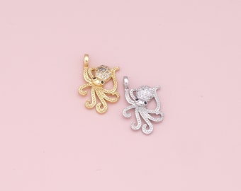 Puede incluir: Dos abalorios de pulpo, uno dorado y otro plateado, con detalles de cristal transparente. Los abalorios están sobre un fondo rosa.