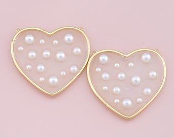 May include: Two gold heart-shaped frames filled with white pearls of varying sizes. The frames are on a pink background.