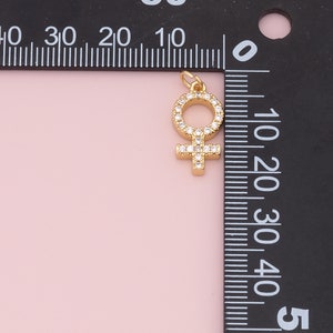 May include: Gold-toned charm featuring a female symbol with clear rhinestones. The charm is measured against a ruler.