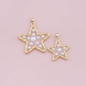 Pu&ograve; includere: Due ciondoli a forma di stella color oro con un bordo strutturato e un centro rosa chiaro. Le stelle sono riempite di perle bianche. I ciondoli sono di diverse dimensioni e sono appesi a un piccolo anello d'oro.