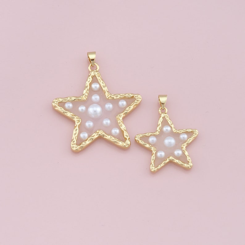 Pu&ograve; includere: Due ciondoli a forma di stella color oro con un bordo strutturato e un centro rosa chiaro. Le stelle sono riempite di perle bianche. I ciondoli sono di diverse dimensioni e sono appesi a un piccolo anello d'oro.