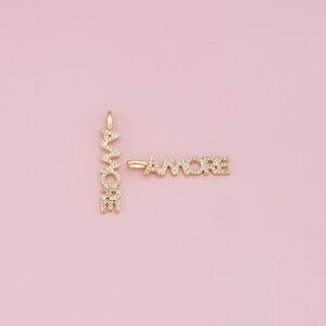May include: A gold charm with the word "amore" spelled out in vertical and horizontal directions, with each letter encrusted with small clear crystals.