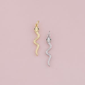 Pode incluir: Dois pingentes de cobra, um dourado e outro prateado, com olhos verdes. Os pingentes estão sobre um fundo branco.