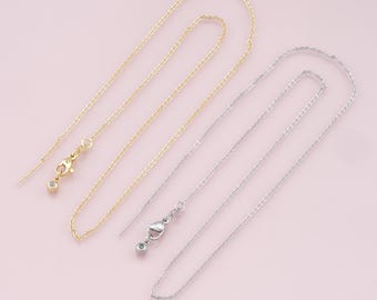 May include: Two delicate chains, one gold and one silver, with small clasps and a tiny bead at the end of each. The chains are laid out on a pink background.