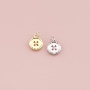 May include: Two button-shaped charms. One is gold-colored with a gold bail, and the other is silver-colored with a silver bail. Both have a cross-shaped design in the center. The background is a solid light pink.