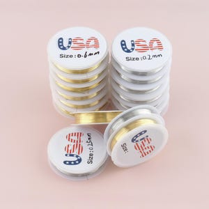 以下が含まれることがあります： 金と銀色のジュエリーワイヤーの巻き取りが積み重ねられています。各巻き取りには、赤と青で「USA」とmm単位のサイズが記載されています。サイズは0.6mm、0.2mm、0.15mmです。