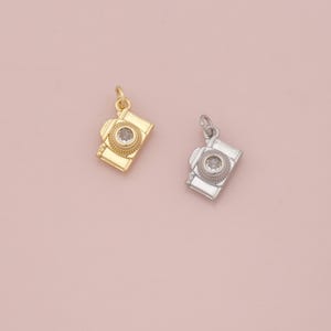Pode incluir: Dois pingentes de câmera, um dourado e outro prateado, são exibidos em um fundo rosa claro. Cada pingente apresenta um design detalhado de câmera com uma gema clara central. Os pingentes são suspensos por pequenos laços.