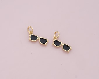 Puede incluir: Dos abalorios de color dorado en forma de gafas de sol con lentes negros. Los abalorios tienen un bucle en la parte superior para sujetarlos a una pulsera o un collar.