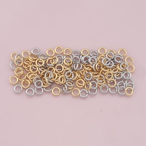 May include: A collection of small, open jump rings in gold and silver tones. These metal rings are scattered across a light pink surface, suitable for jewelry making and crafting projects.