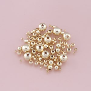 May include: A large group of gold-colored metal beads of various sizes. The beads are scattered on a pink surface.