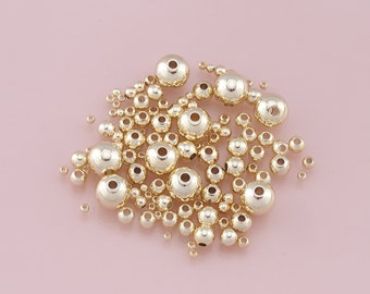 May include: A large group of gold-colored metal beads of various sizes. The beads are scattered on a pink surface.