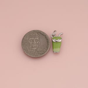 May include: A silver-toned metal charm shaped like a cup with a straw, featuring light green enamel detailing. The charm is positioned next to a United States nickel coin for scale, showing the relative size of the small beverage-themed jewelry piece.