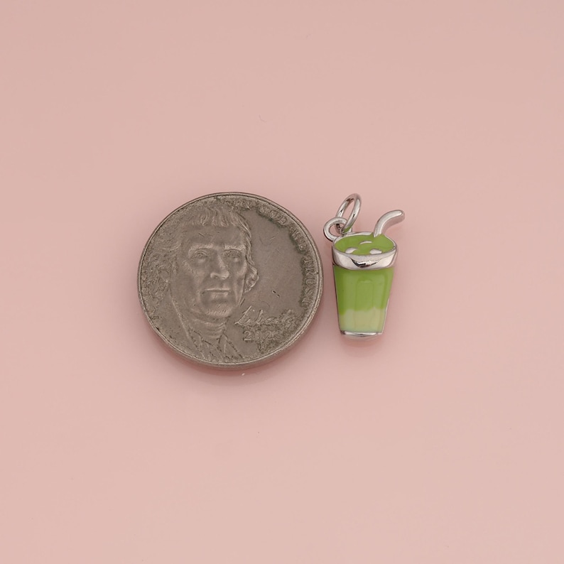 May include: A silver-toned metal charm shaped like a cup with a straw, featuring light green enamel detailing. The charm is positioned next to a United States nickel coin for scale, showing the relative size of the small beverage-themed jewelry piece.