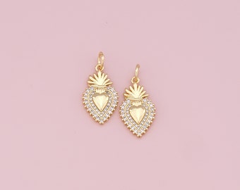 May include: Two gold-tone heart-shaped charms with a detailed design and clear crystals. The charms are on a pink background.