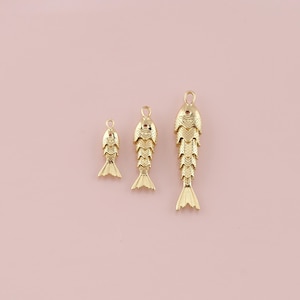 May include: Three gold-coloured fish charms of varying sizes are arranged against a soft pink background. Each charm features a detailed fish design with scales and a tail fin. The charms have a loop at the top for attaching to jewellery.