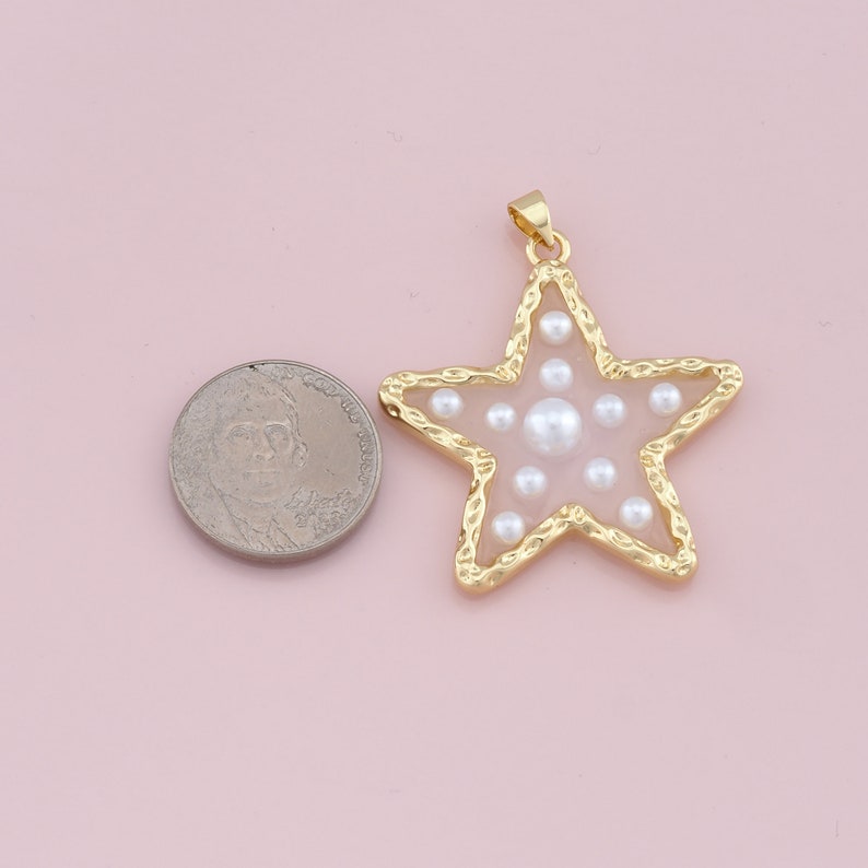 Pu&ograve; includere: Ciondolo a forma di stella color oro con bordo strutturato, riempito di perle bianche. Il ciondolo &egrave; accanto a una moneta per il confronto delle dimensioni. La stella ha un piccolo anello nella parte superiore.