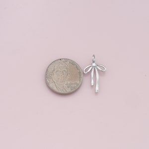 May include: Silver charm in the shape of a bow with a clear rhinestone in the center. The charm is about the size of a US quarter.