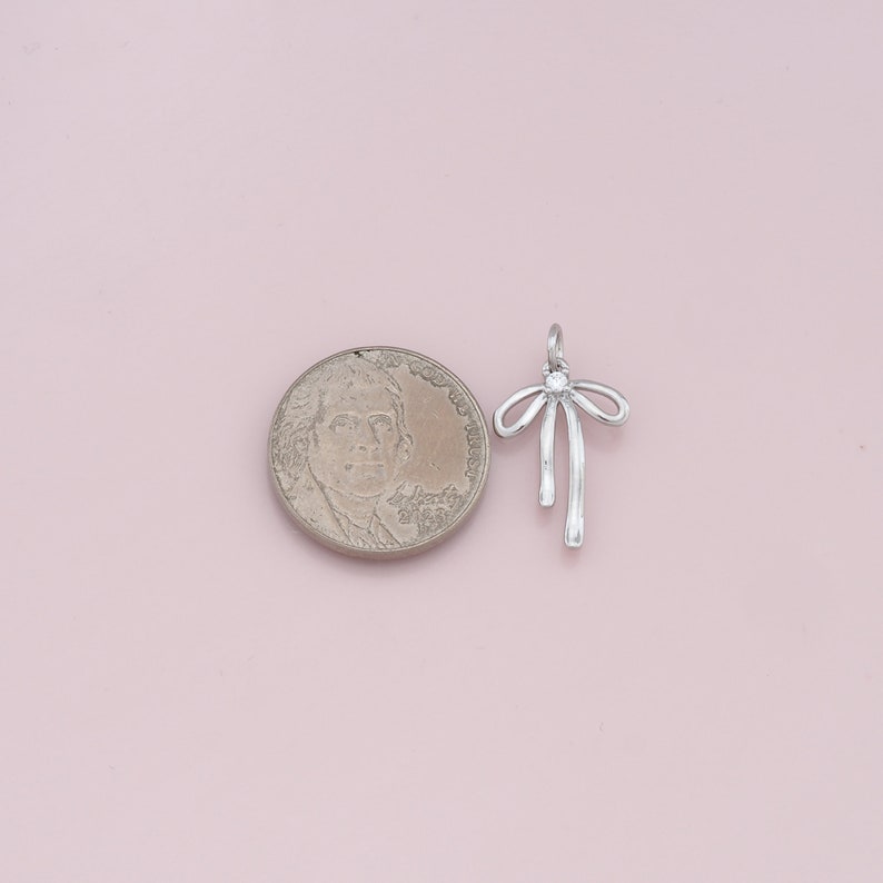 May include: Silver charm in the shape of a bow with a clear rhinestone in the center. The charm is about the size of a US quarter.