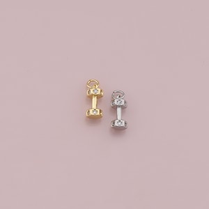 May include: Two dumbbell charms, one gold-colored and one silver-colored, are displayed against a pink background. Each charm features a barbell design with clear, sparkling accents. The charms are small and detailed, suitable for jewelry making.