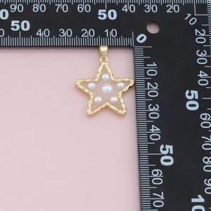 Pu&ograve; includere: Ciondolo a forma di stella color oro con centro rosa chiaro e perle bianche incastonate. Il ciondolo ha un bordo dorato strutturato e un piccolo anello per una catena. Lo sfondo &egrave; rosa chiaro.