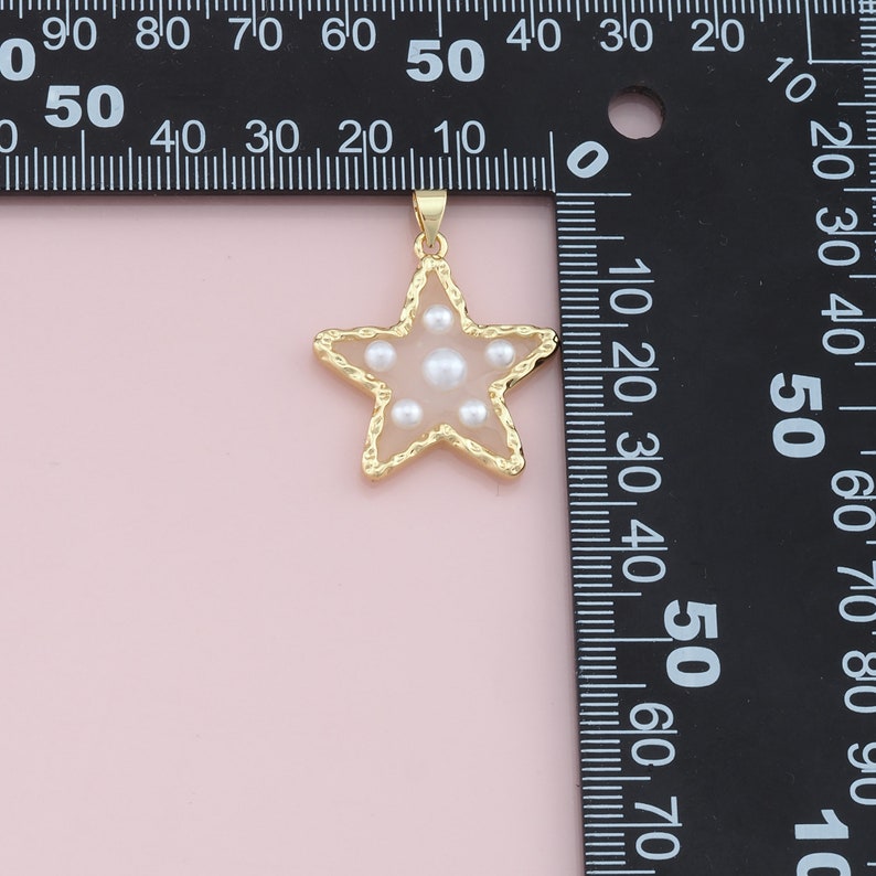 Pu&ograve; includere: Ciondolo a forma di stella color oro con centro rosa chiaro e perle bianche incastonate. Il ciondolo ha un bordo dorato strutturato e un piccolo anello per una catena. Lo sfondo &egrave; rosa chiaro.