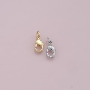 May include: Two lobster claw clasps, one gold and one silver, on a pink background.