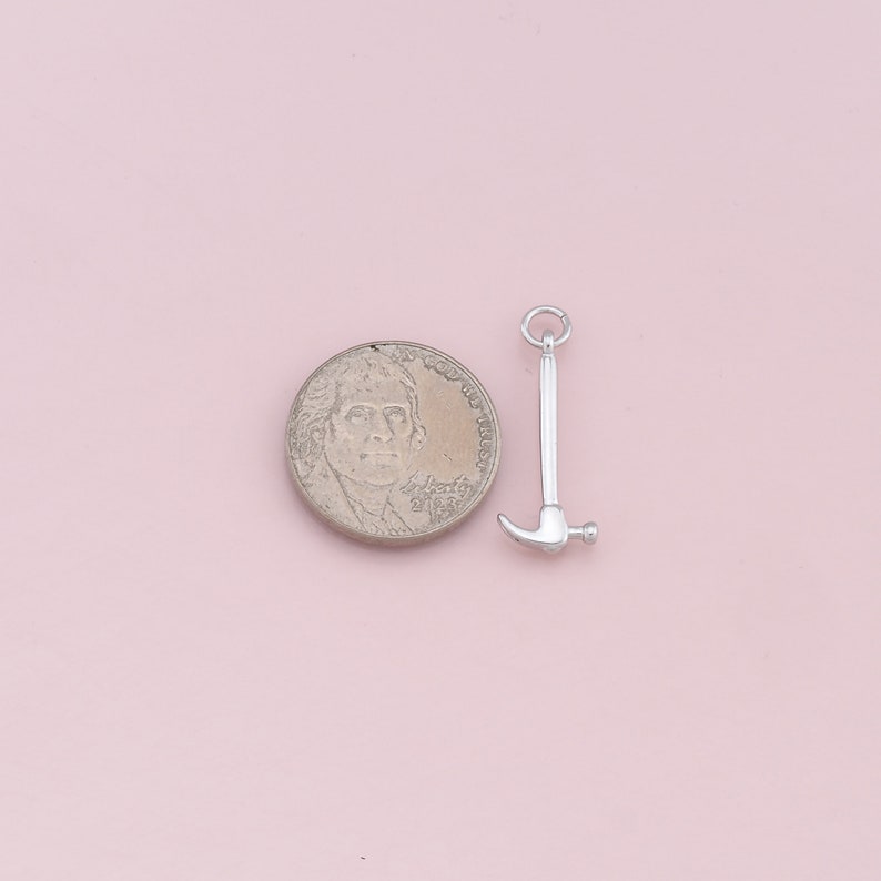 May include: Silver charm in the shape of a hammer, next to a US penny for size comparison.