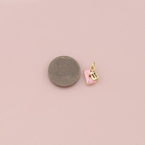 May include: A gold-colored charm featuring a pink envelope with two red hearts and a small paper with the words "I love you". The charm is likely for a necklace or bracelet, and is on a pink background.