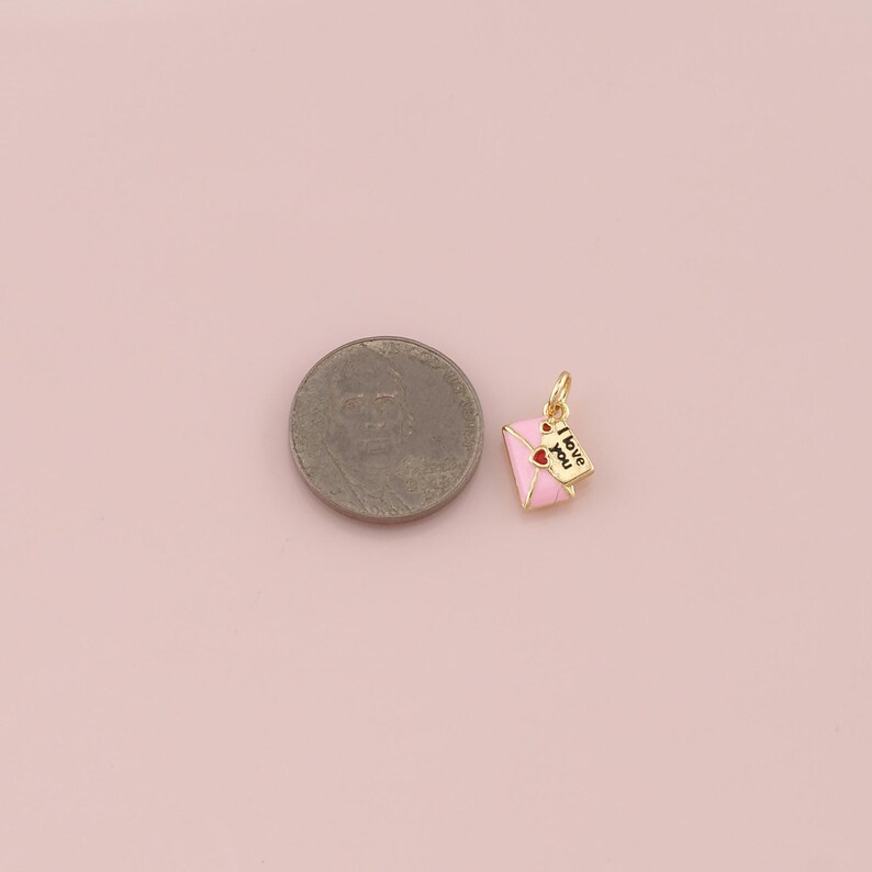 May include: A gold-colored charm featuring a pink envelope with two red hearts and a small paper with the words "I love you". The charm is likely for a necklace or bracelet, and is on a pink background.
