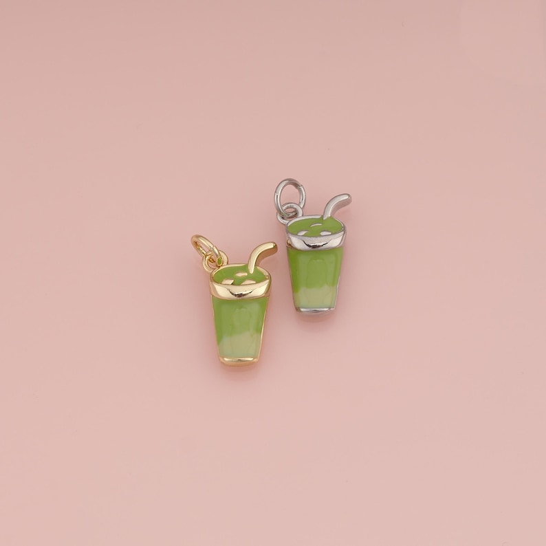 May include: Two small charms shaped like drinking glasses filled with a green beverage. One charm is gold-toned with a light green enamel fill, and the other is silver-toned with a similar green enamel design, both featuring a small straw.