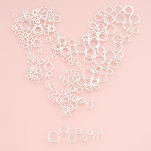 May include: A collection of numerous silver-colored metal jump rings of varying diameters arranged in a heart shape, with a small row of individual jump rings of different sizes positioned separately below the main heart-shaped arrangement.